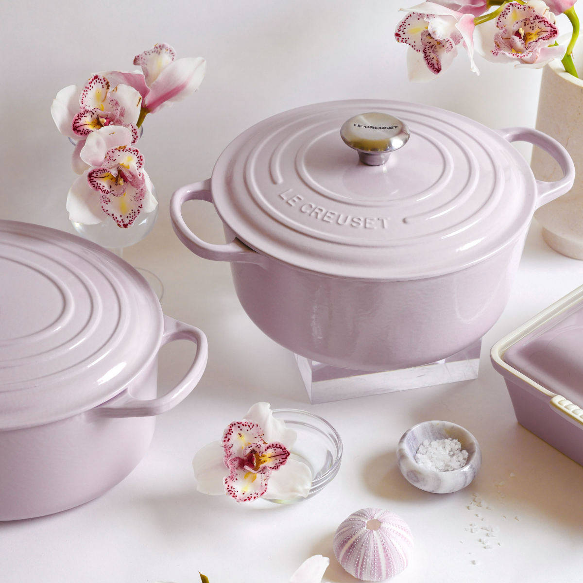 Overhead of Le Creuset Shallot casserole with pink flowers and glass bowls on a pastel background.