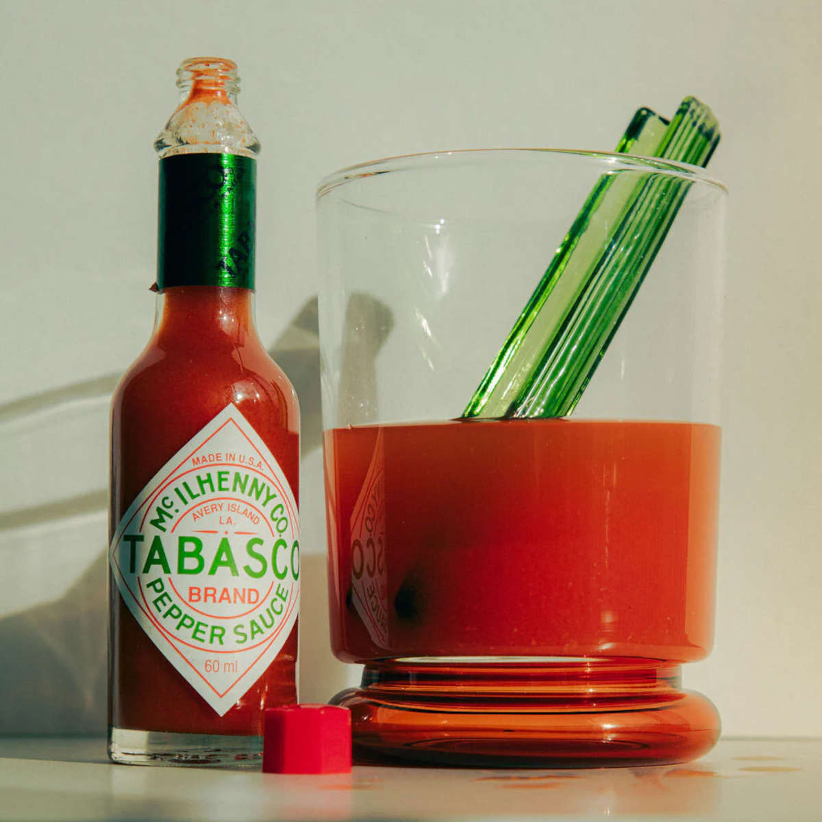 Maison Balzac Bloody Mary Cocktail Glass – clear glass with red base, filled with tomato juice, green glass stirrer placed beside on white surface