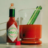 Maison Balzac Bloody Mary Cocktail Glass – clear glass with red base, filled with tomato juice, green glass stirrer placed beside on white surface