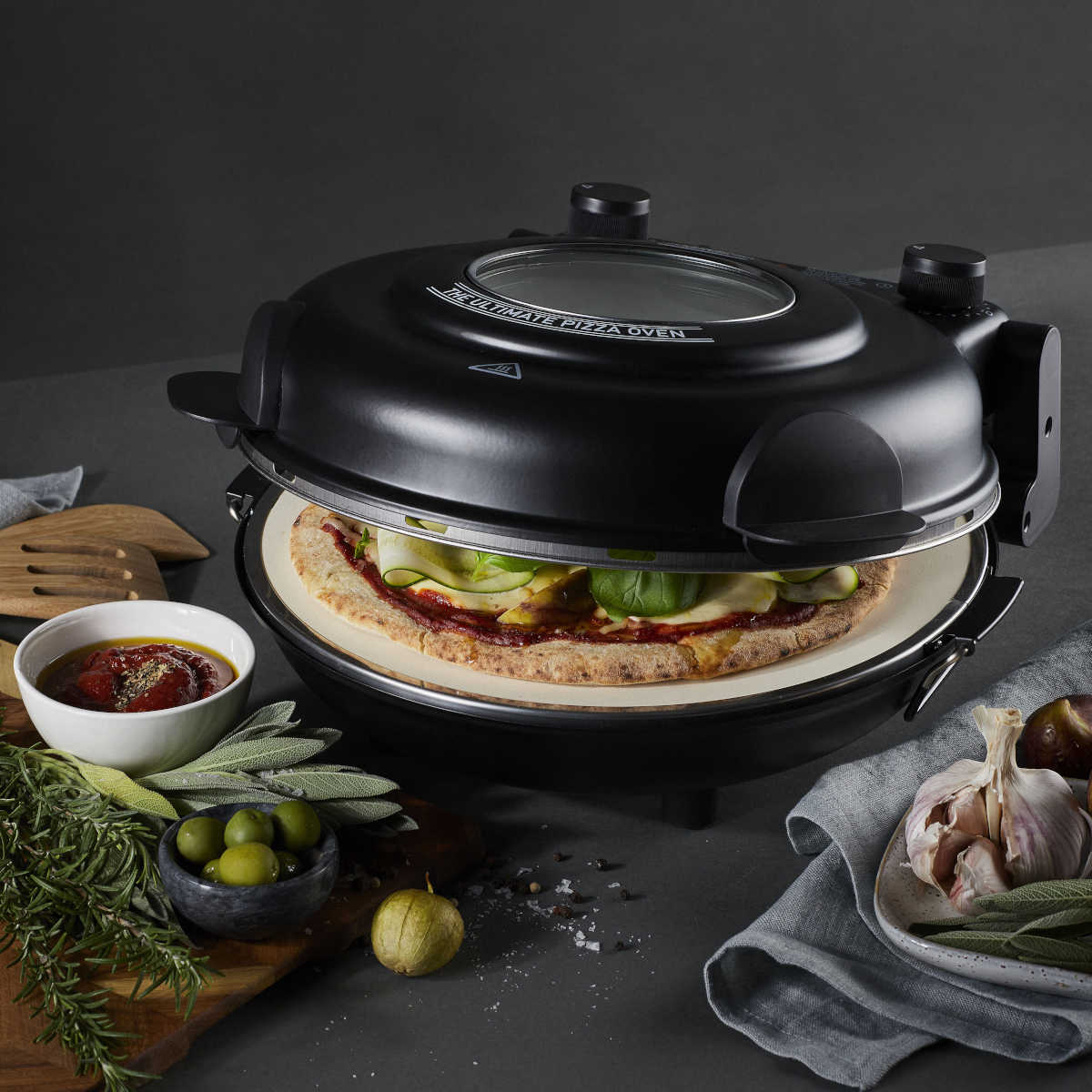 Angled view of MasterPro Ultimate Pizza Oven Black baking pizza with herbs and sauce nearby
