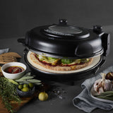 Angled view of MasterPro Ultimate Pizza Oven Black baking pizza with herbs and sauce nearby