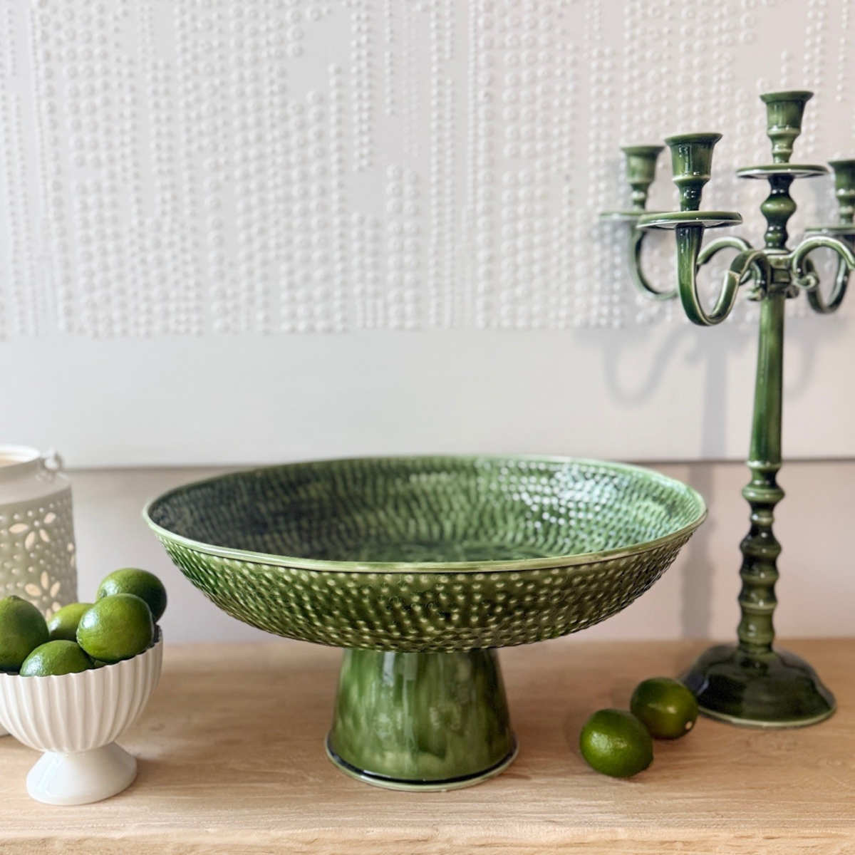 Mediterranean Market Wash Enamel Footed Tray Green 47cm, lifestyle shot on wooden table with limes and green candle holder against textured wall